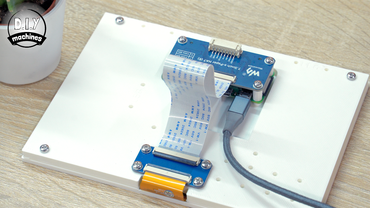 A white display screen connected to a blue driver board via a flat ribbon cable, resting on a light-colored table.