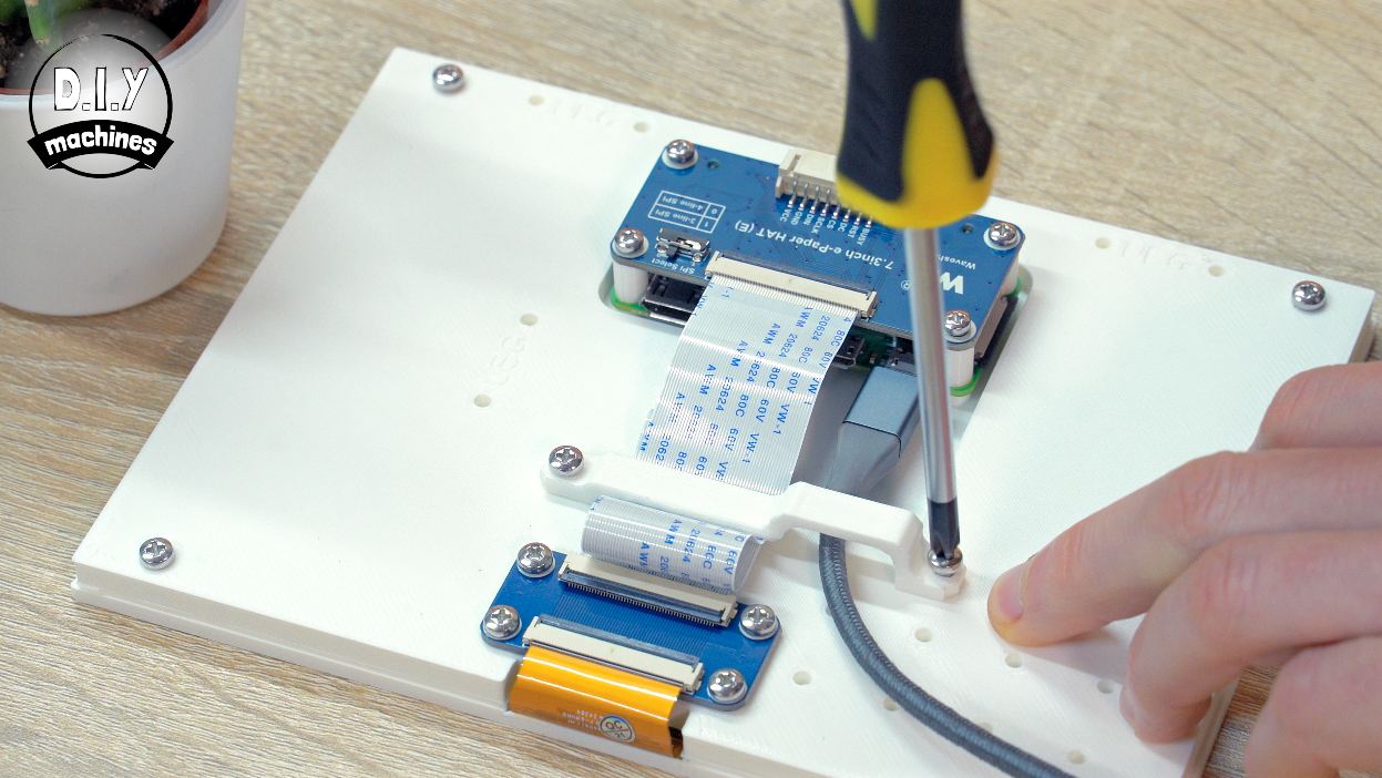A hand uses a yellow-handled screwdriver to mount blue electronic components onto a white board with ribbon cables.