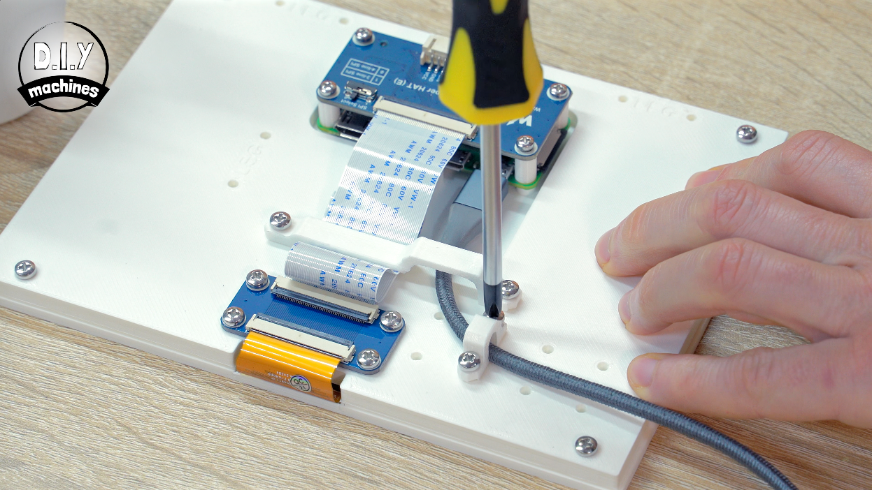 A person uses a screwdriver to secure a grey cable clamp onto a white 3D-printed panel with mounted electronic components.