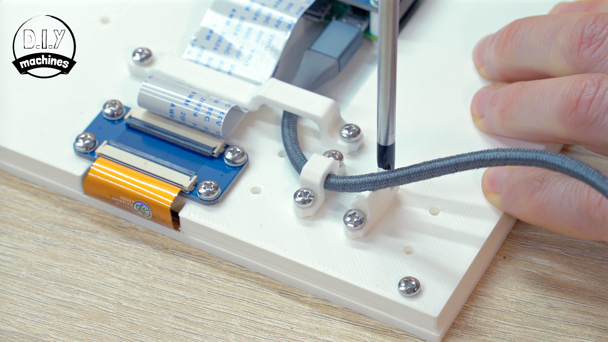 A screwdriver secures a cable clamp onto a white mounting board next to electronic components and a ribbon cable.
