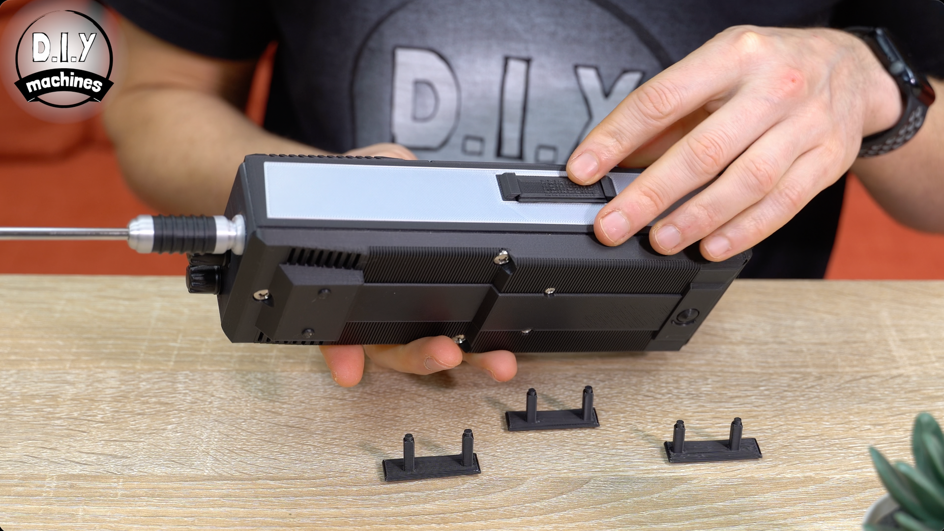 A person holds a black rectangular device with an antenna, while small plastic components sit on a wooden table.
