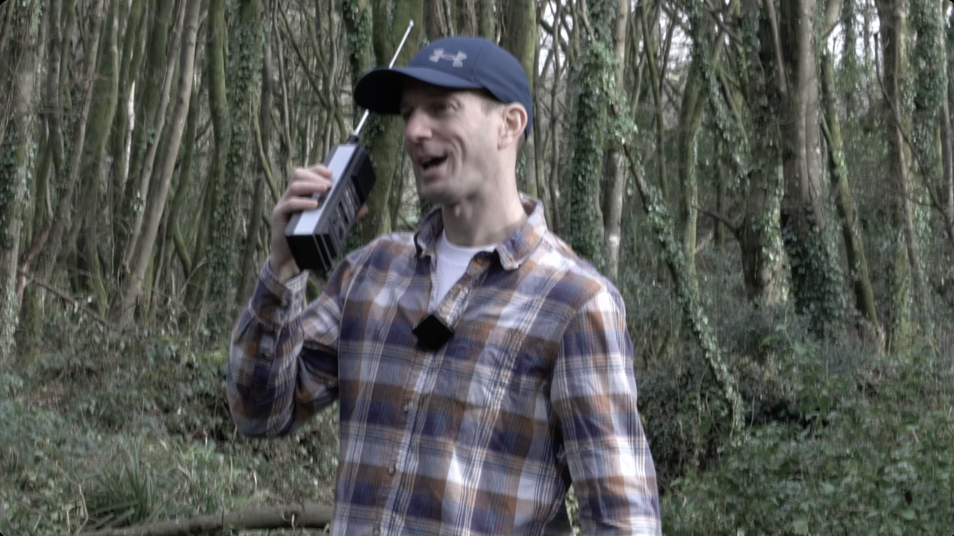 A person in a plaid shirt and baseball cap speaks into a walkie-talkie while standing in a wooded area.