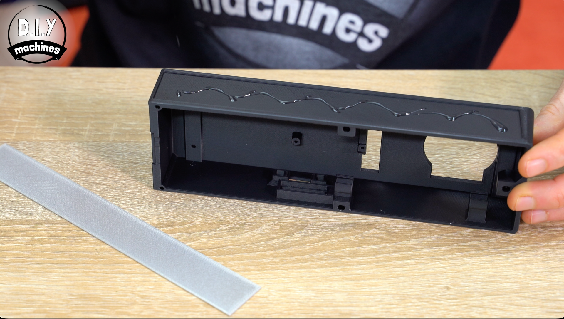 A black 3D-printed plastic component held by a hand next to a sanding tool on a wooden surface.