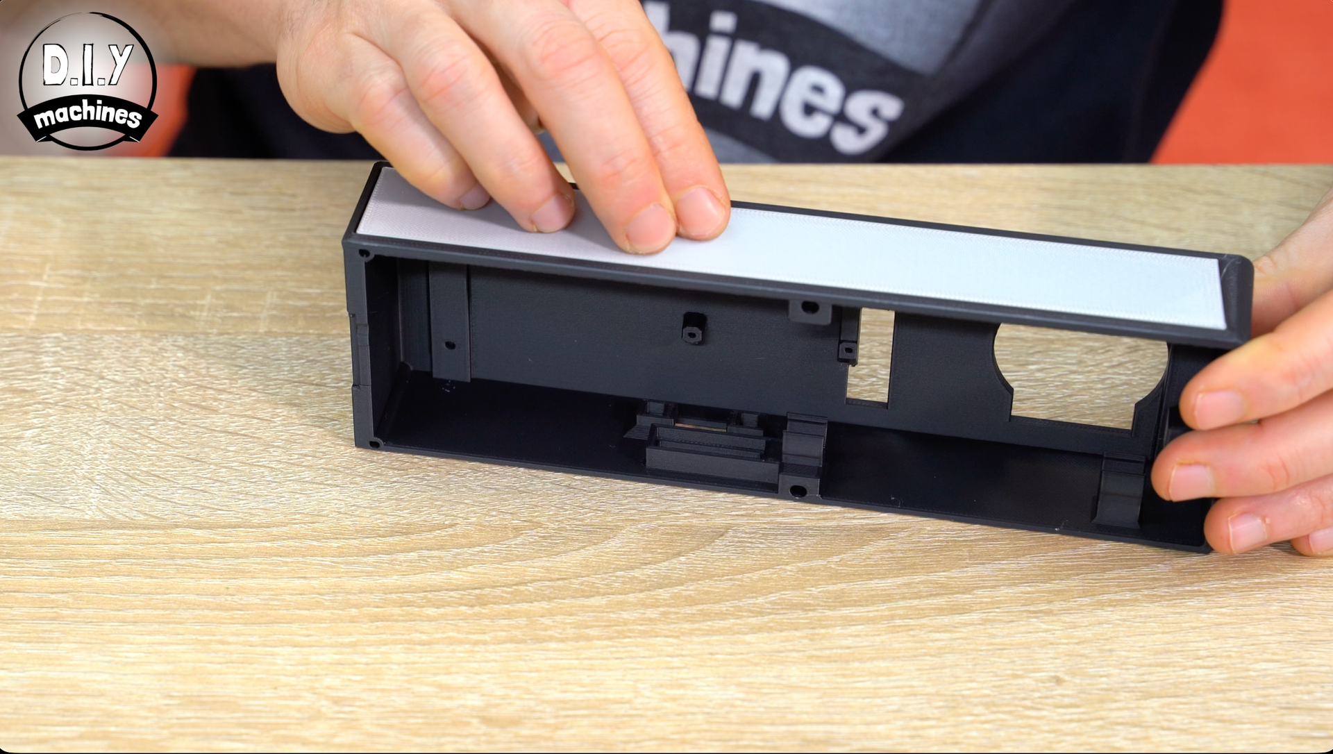 A person aligns a white adhesive strip onto the top edge of a black 3D-printed plastic housing on a wooden surface.