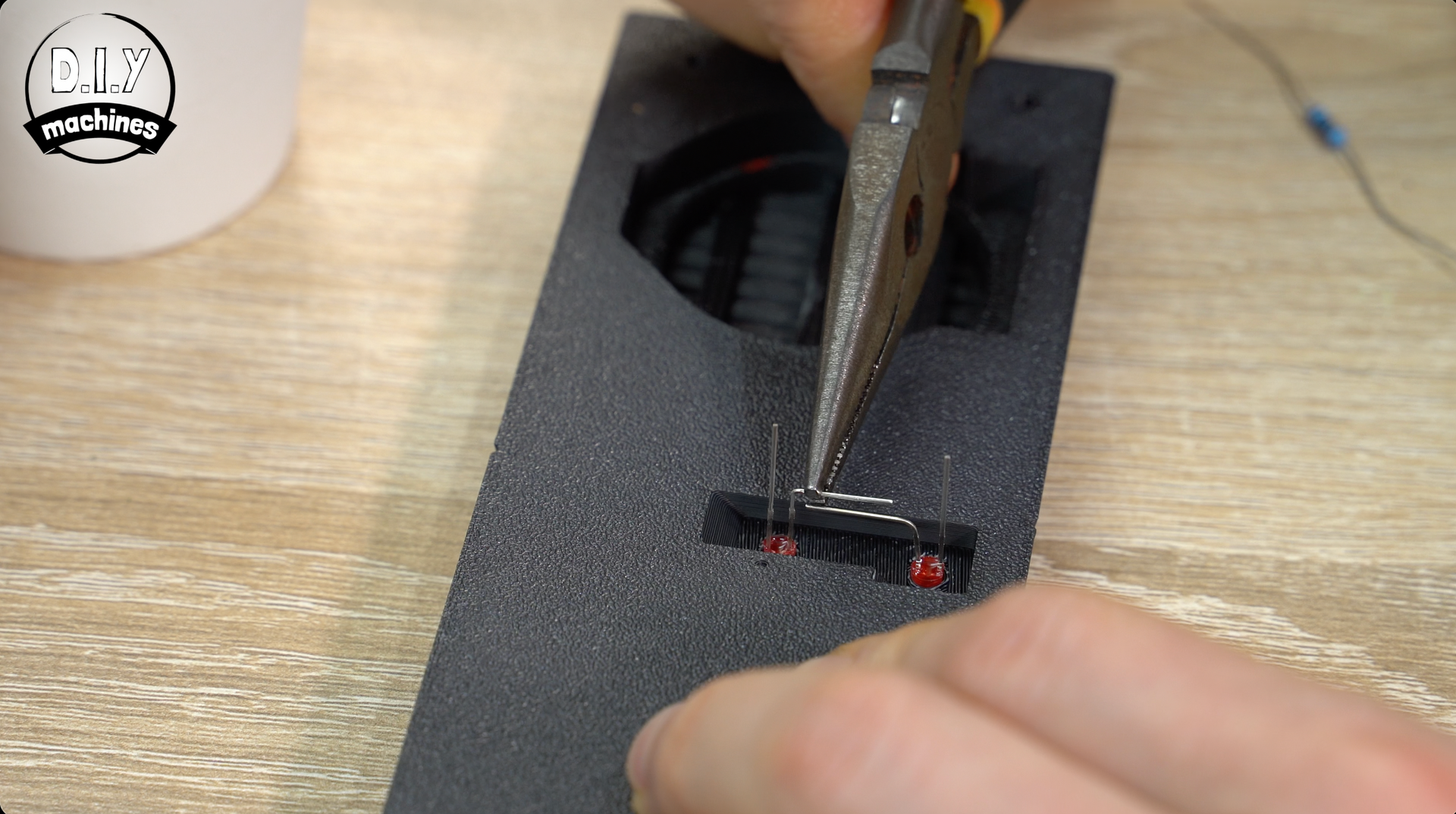 A person uses needle-nose pliers to insert small electronic components into a black foam block on a wooden surface.