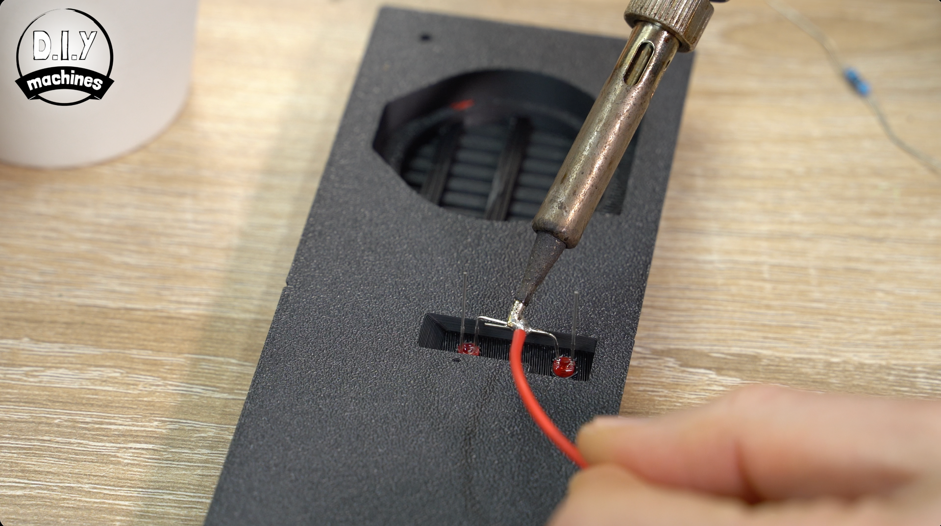 A person uses a soldering iron to attach a red wire to a component on a textured black electronic housing.