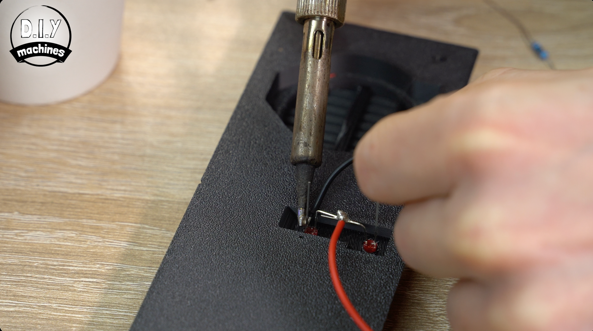 A person uses a soldering iron to attach a red wire to a small electronic component on a textured black surface.