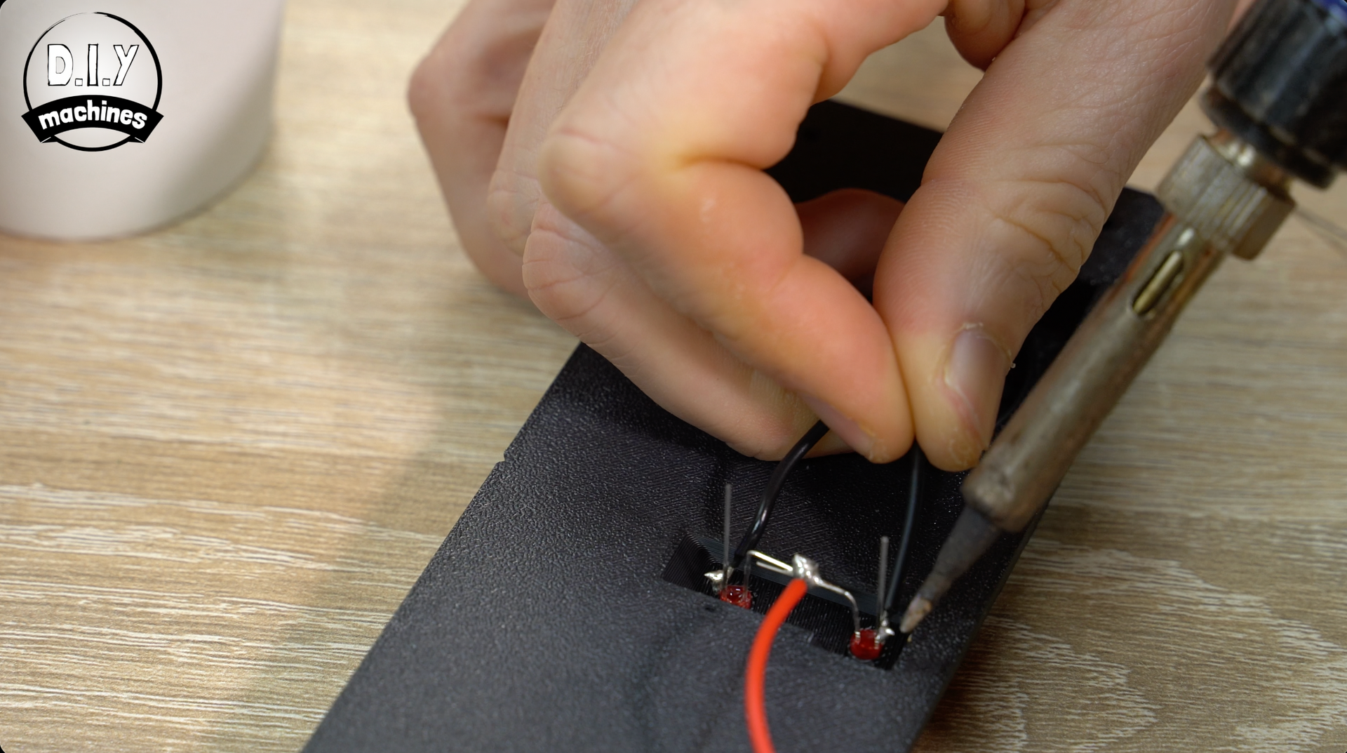 A person uses a soldering iron to attach a wire to a small electronic component embedded in a black foam base.
