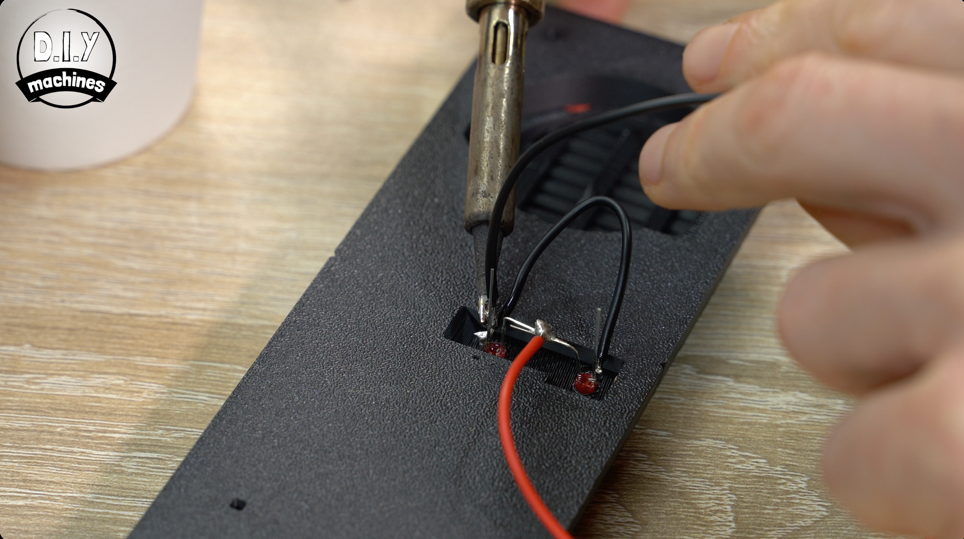 A person uses a soldering iron to attach a black wire to a small circuit board component on a black plastic surface.