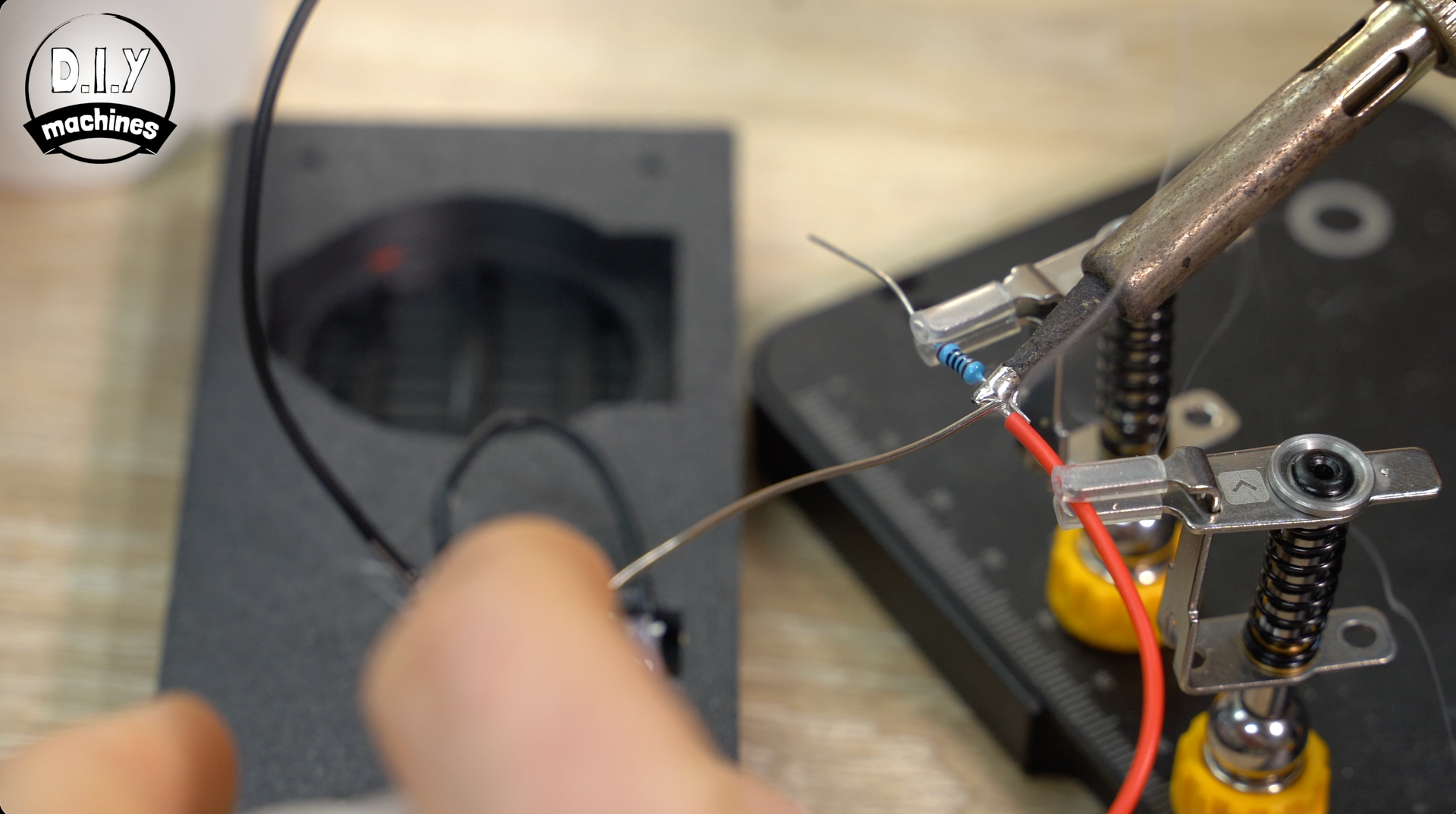 A close-up of a person soldering a red wire to a small blue electronic component held by a helping hands tool.