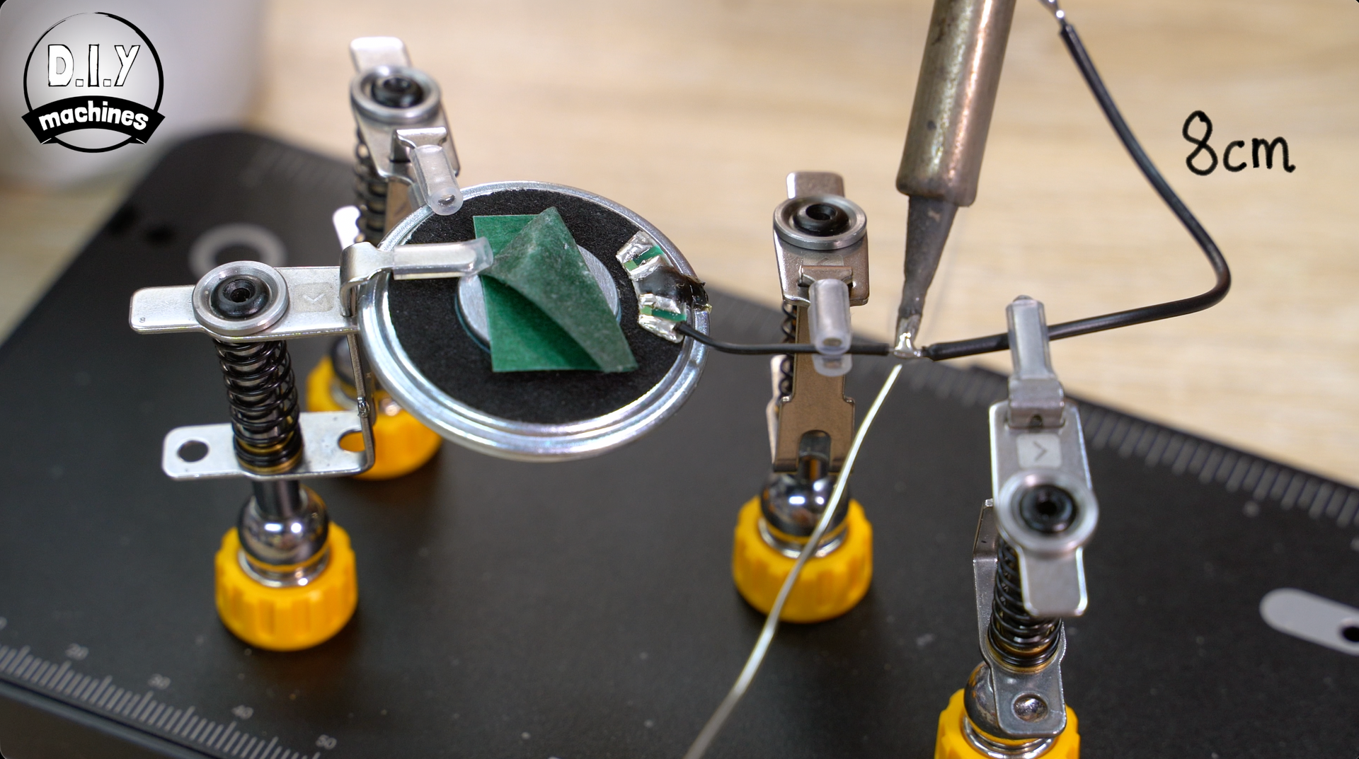 A soldering iron joins a wire to a small circuit board, held in place by a multi-armed workbench with yellow knobs.