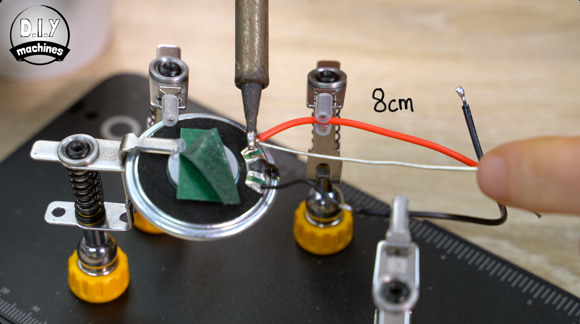 A soldering iron joins a red wire to a speaker terminal held by a helping hand tool on a workstation mat.