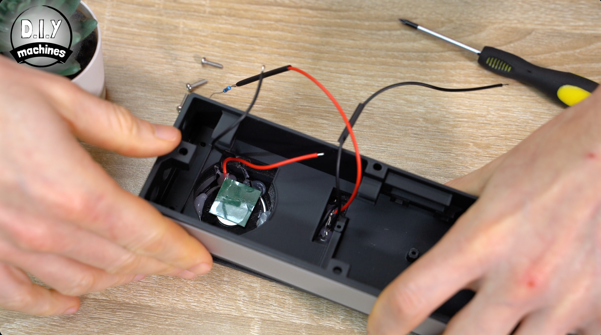 Hands assembling a small electronics device inside a black casing, with wires, a circuit board, and a screwdriver nearby.