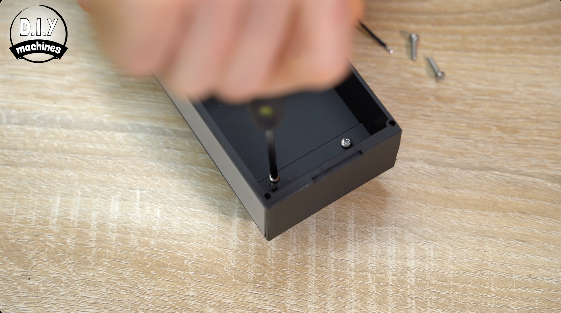 A hand uses a screwdriver to tighten a screw into a black rectangular case on a light wood surface.