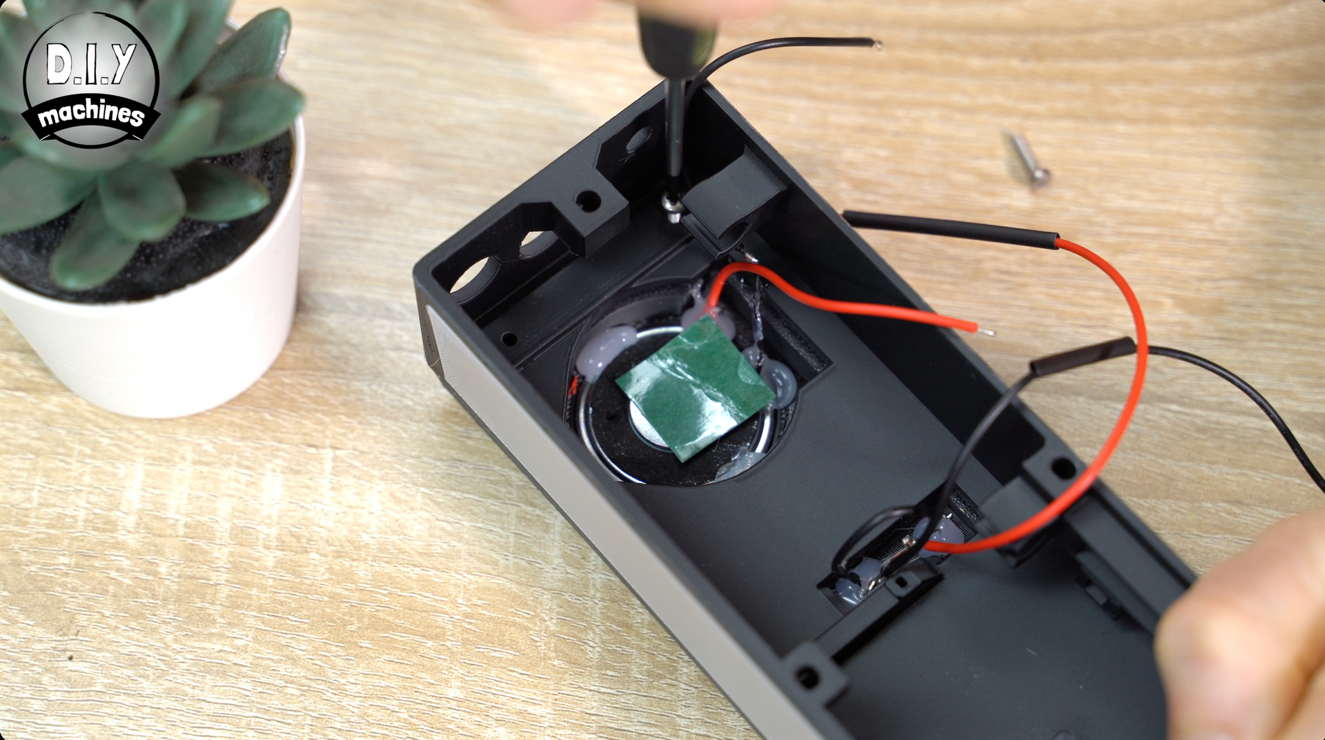 A close-up of a person using a screwdriver to assemble a small black electronic device with red and black wires.