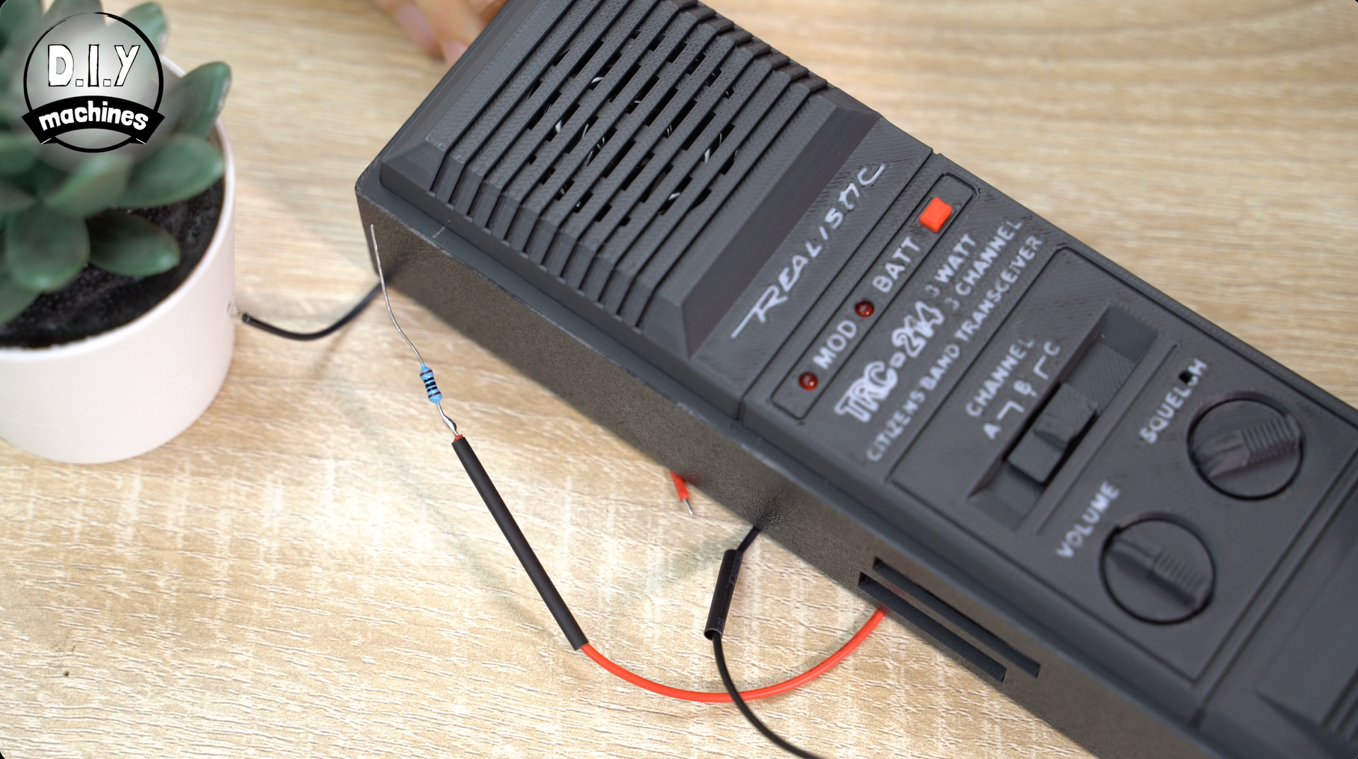 A black Realistic walkie-talkie on a wooden surface with wires and a resistor connected to it next to a small potted plant.