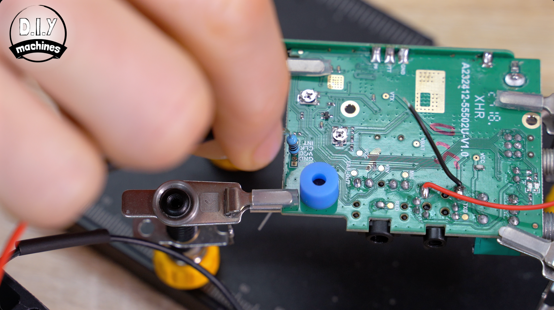 A person uses a helping hand tool to hold a green circuit board while soldering a wire onto its surface.