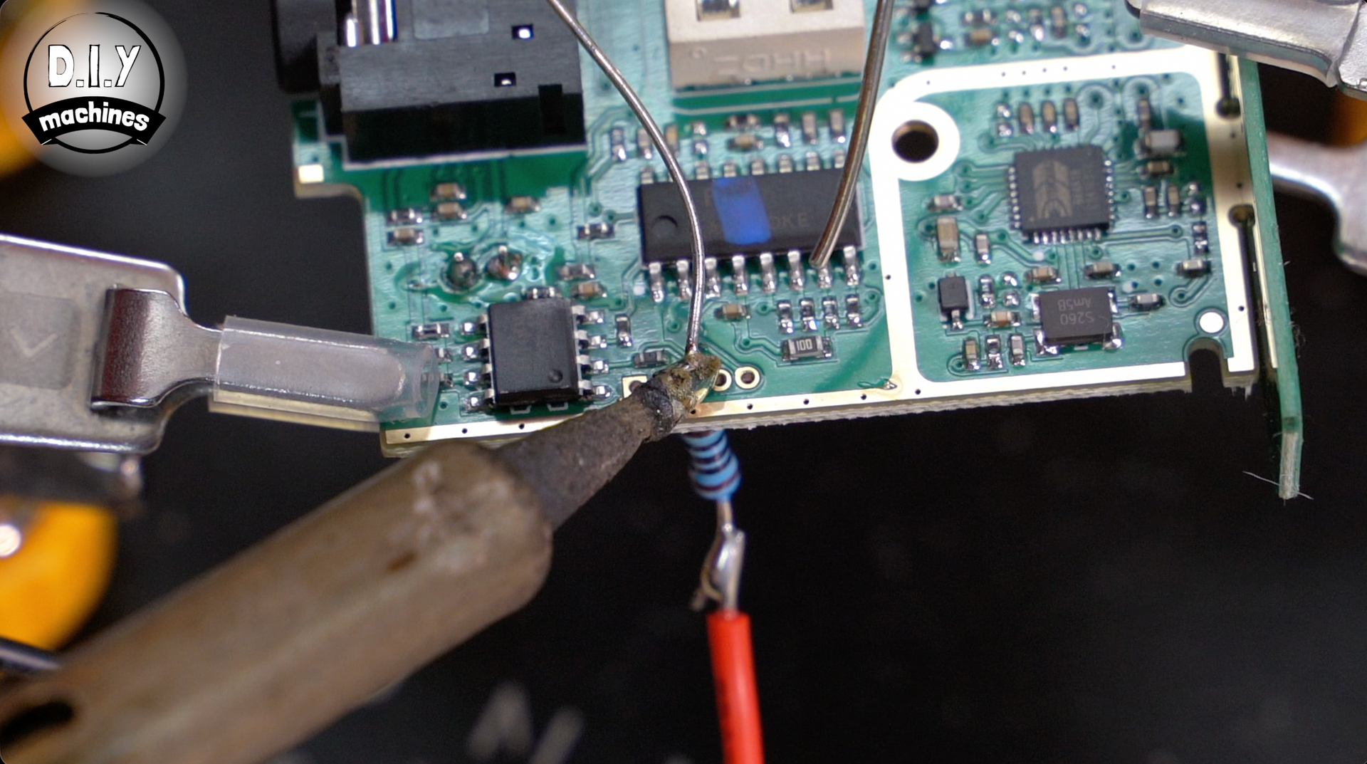 A soldering iron applies heat to a wire connection on an electronic circuit board held by metal clamps.