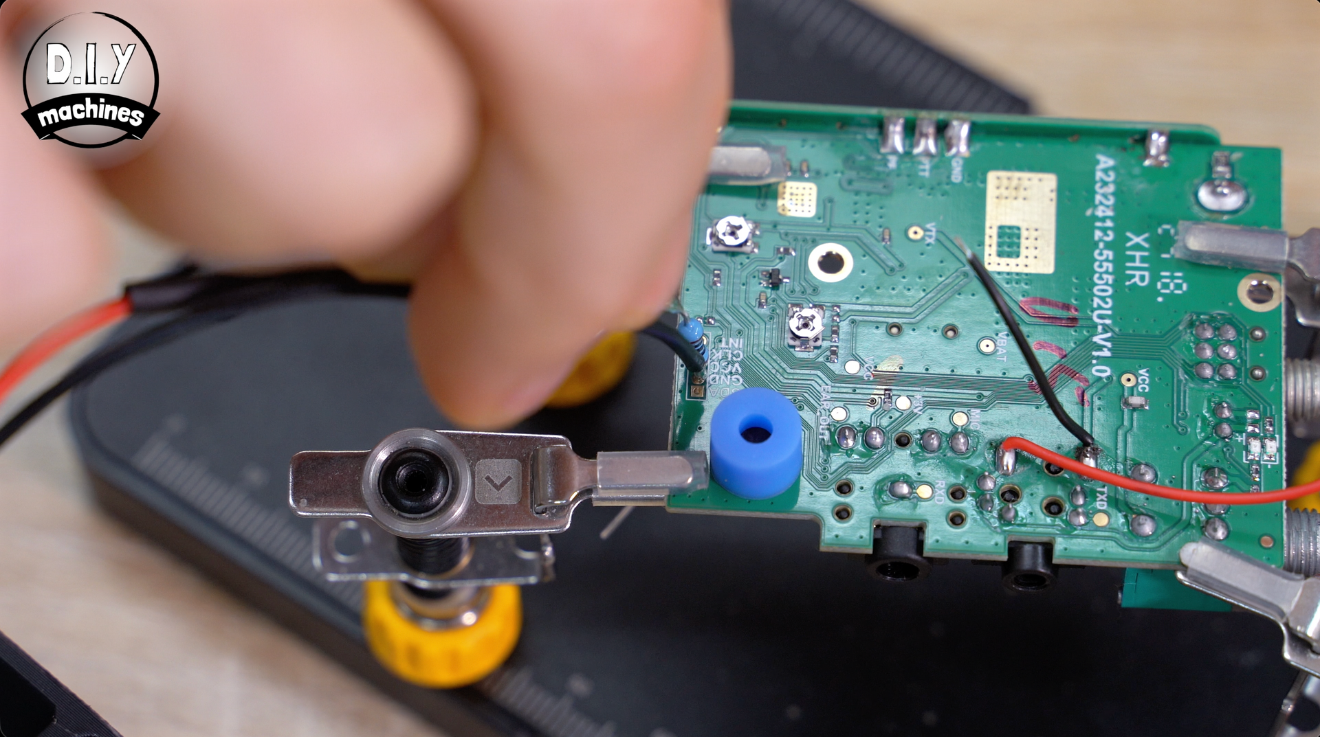 A person holds a black wire against a green circuit board secured in a soldering stand with yellow adjustment knobs.