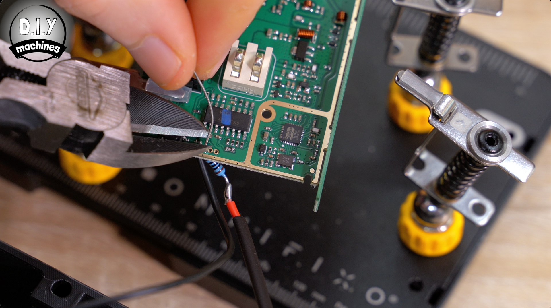 A hand uses pliers to hold a wire soldered to a resistor on a green circuit board held by an electronics repair stand.