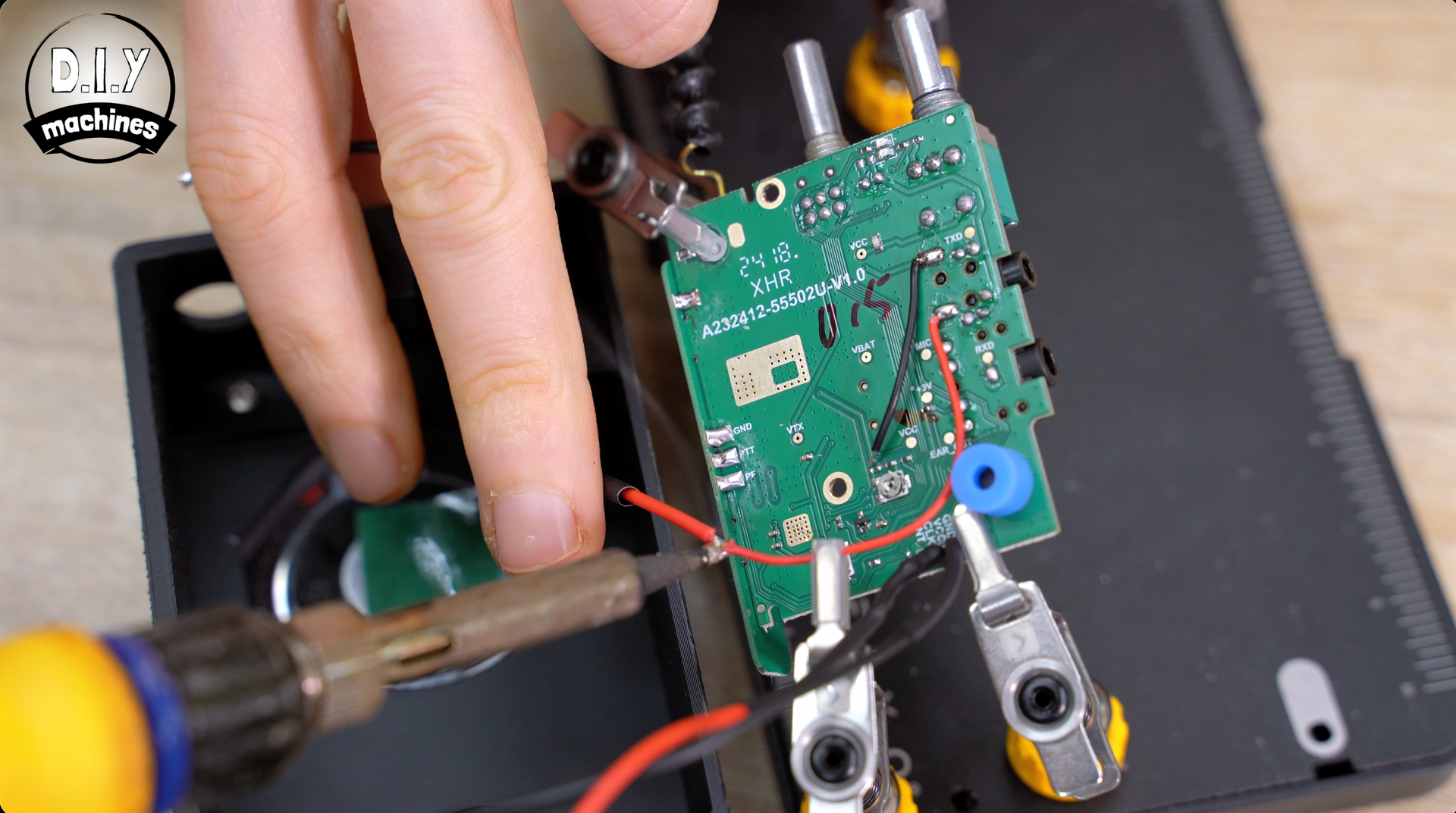A soldering iron tip is melting solder onto a red wire connected to a small green circuit board held by a helping hand tool.