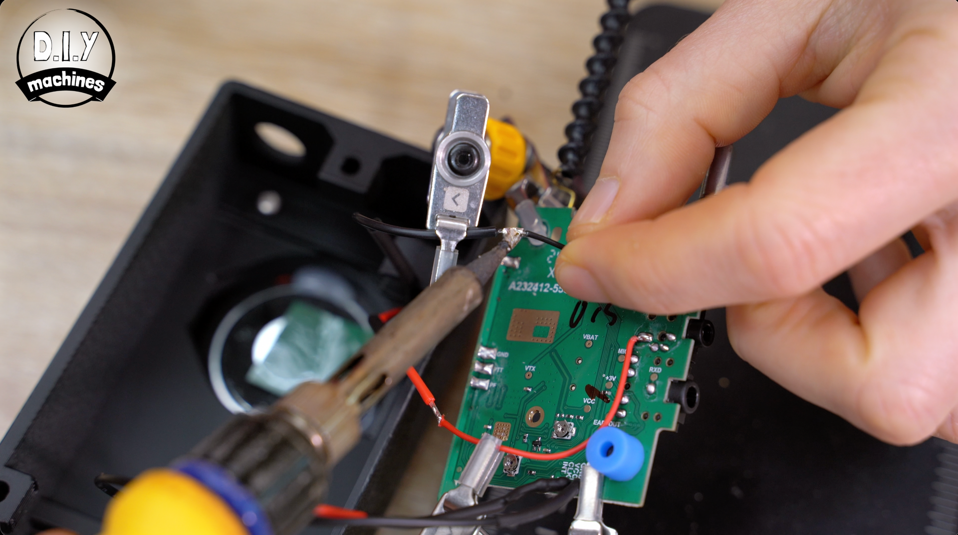 A person uses a soldering iron to attach a wire to a small green circuit board inside a black electronic housing.