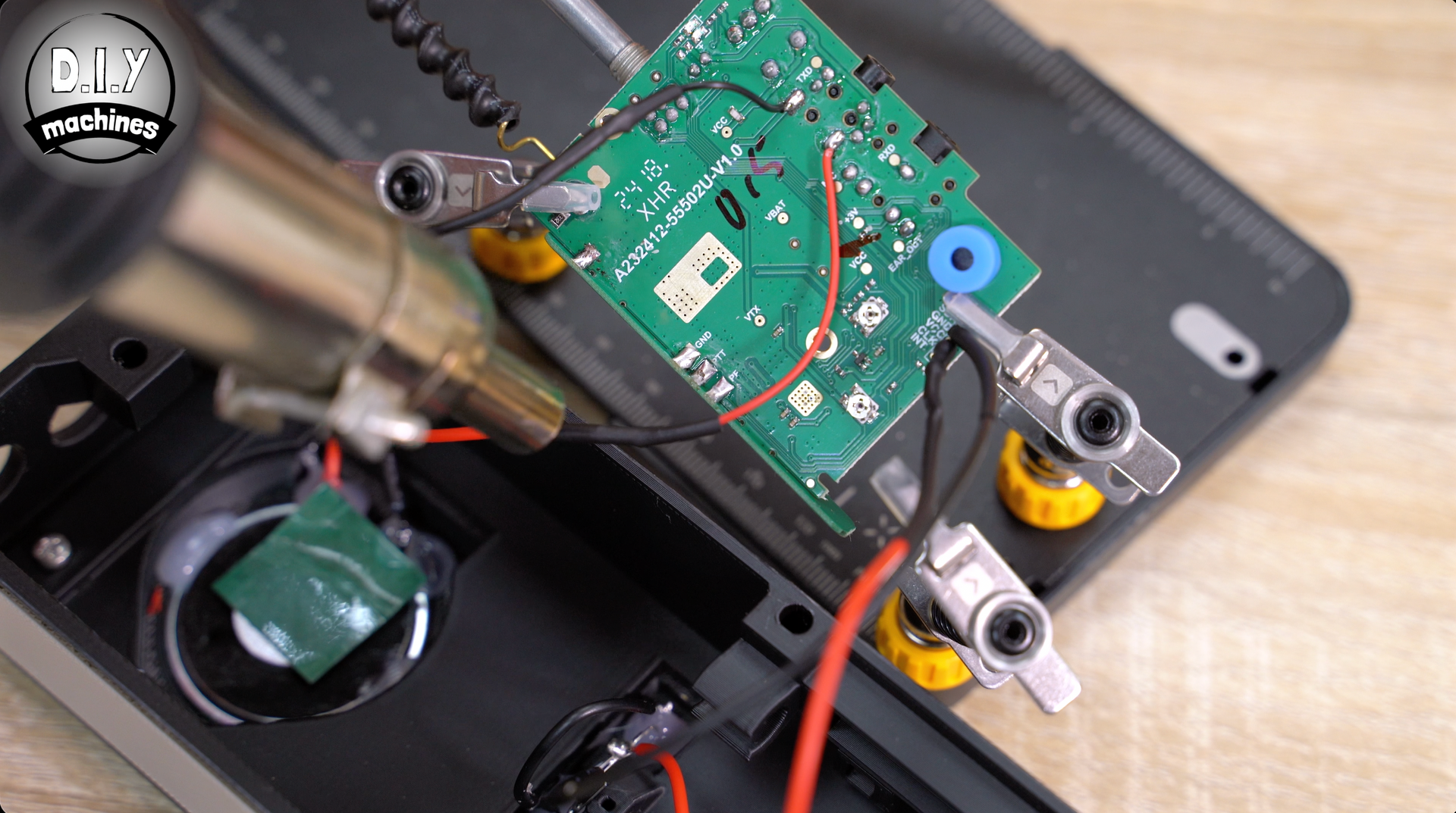 A hot air rework station nozzle directs heat onto a green circuit board held by flexible metal clamps.