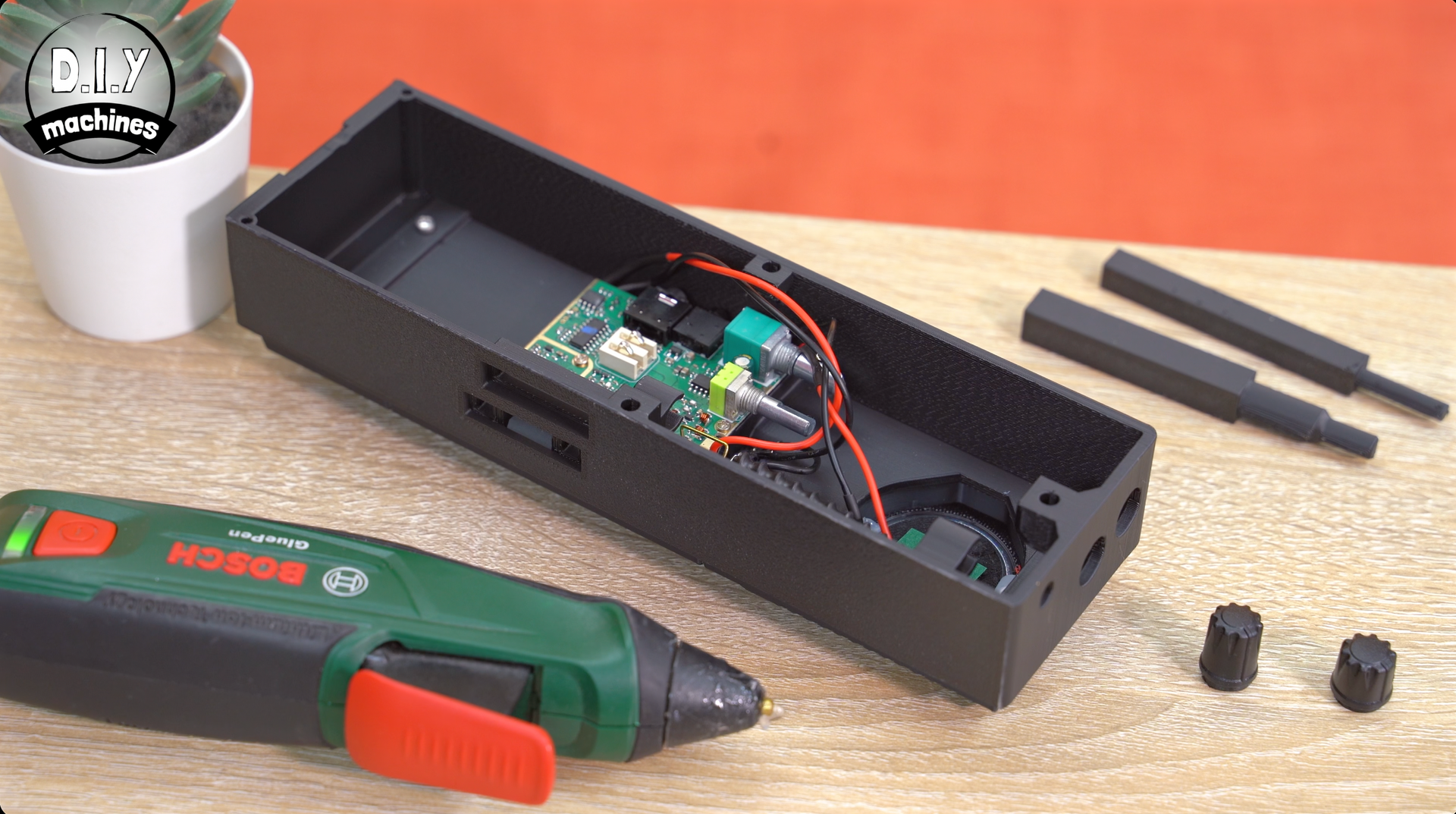 An open black plastic electronics enclosure with internal circuitry, next to a Bosch power tool and two small knobs.