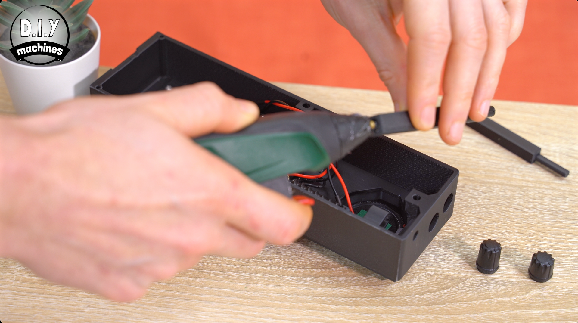 Hands use a hot glue gun to attach a small plastic piece inside a black rectangular box on a wooden desk.