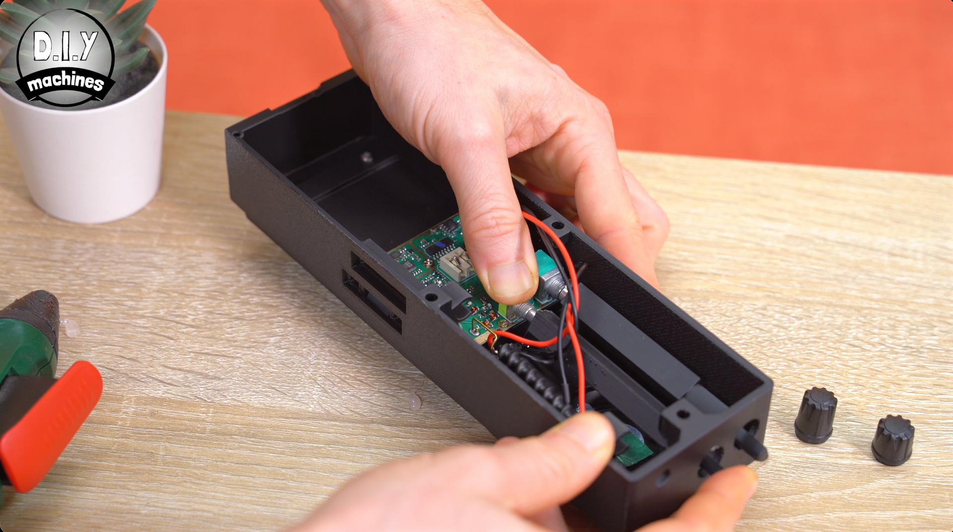 Hands place a circuit board inside a black rectangular plastic housing on a wooden desk with two small knobs nearby.
