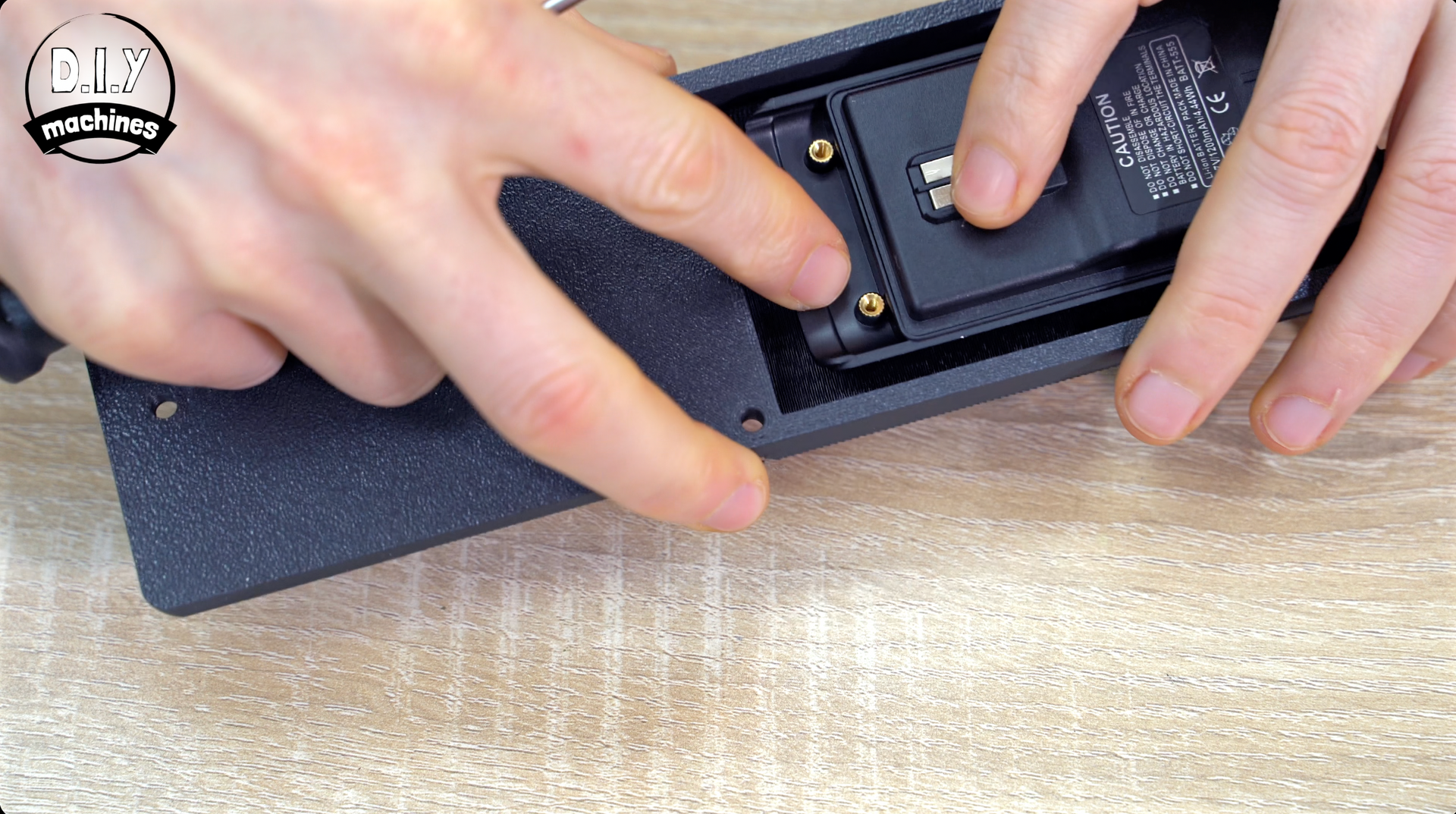 Hands hold a black rectangular electronic component with gold screws while assembling it into a textured device housing.