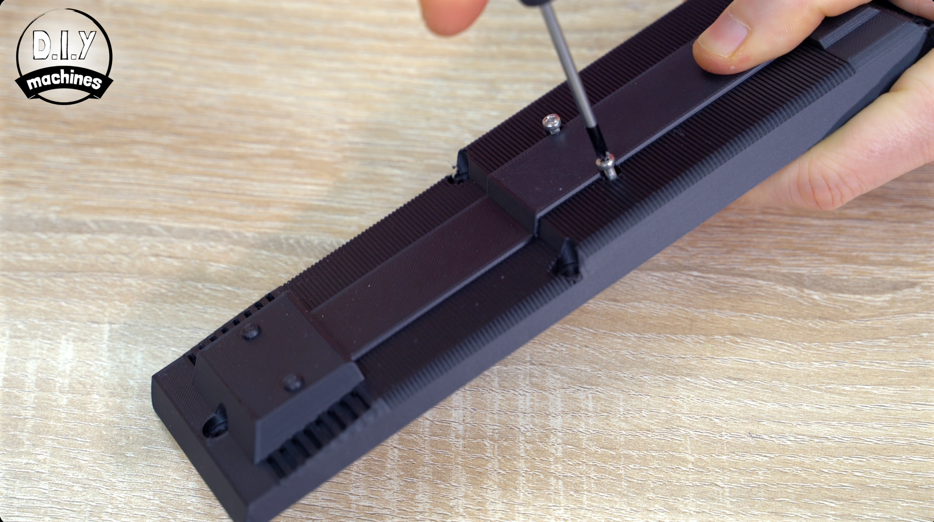 A hand uses a screwdriver to tighten a screw into a black plastic rectangular component on a wooden surface.
