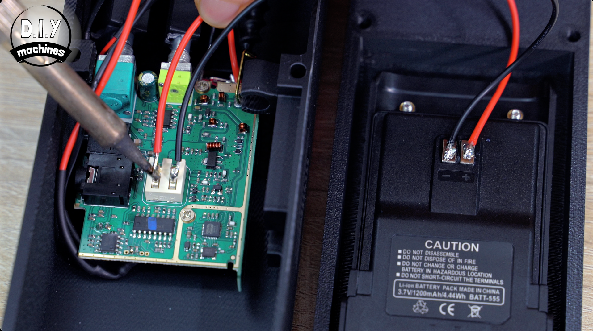 A soldering iron works on a circuit board with two wires attached, positioned next to a device casing with a battery area.