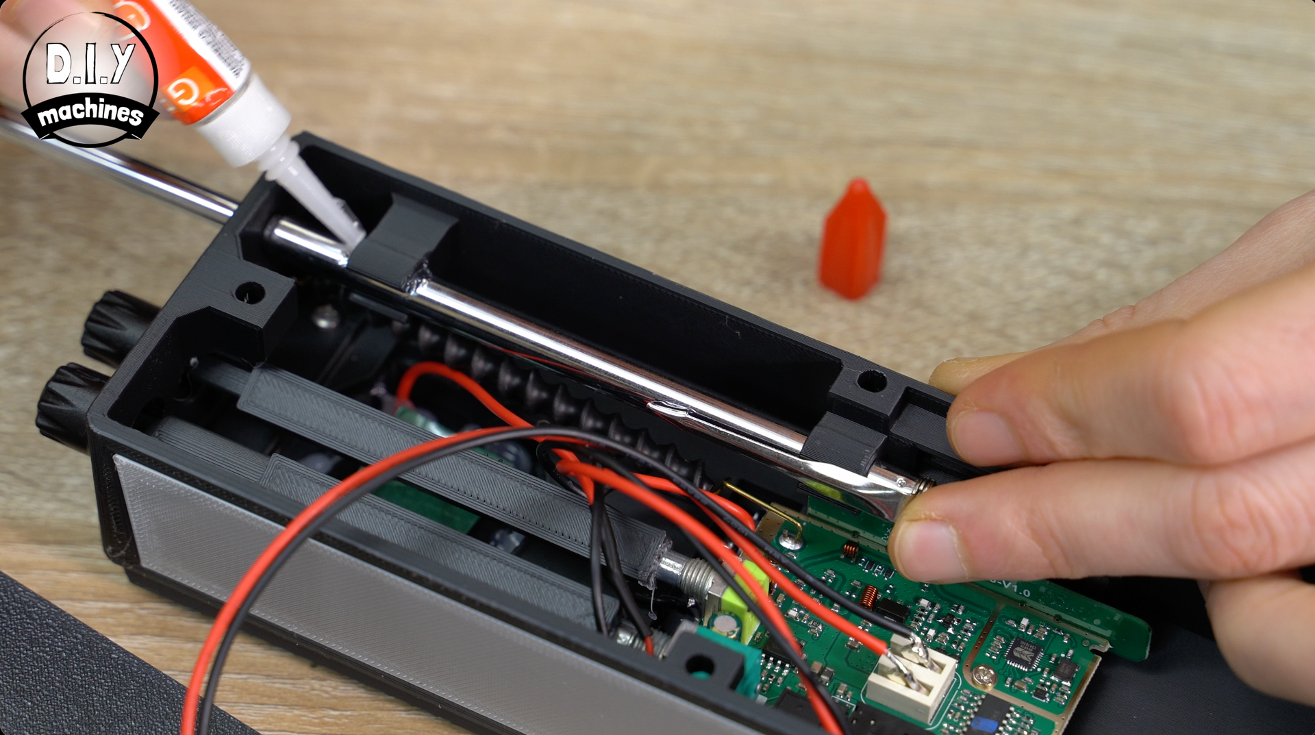 Hands applying glue to a metal rod inside a black 3D-printed enclosure containing electronic components.