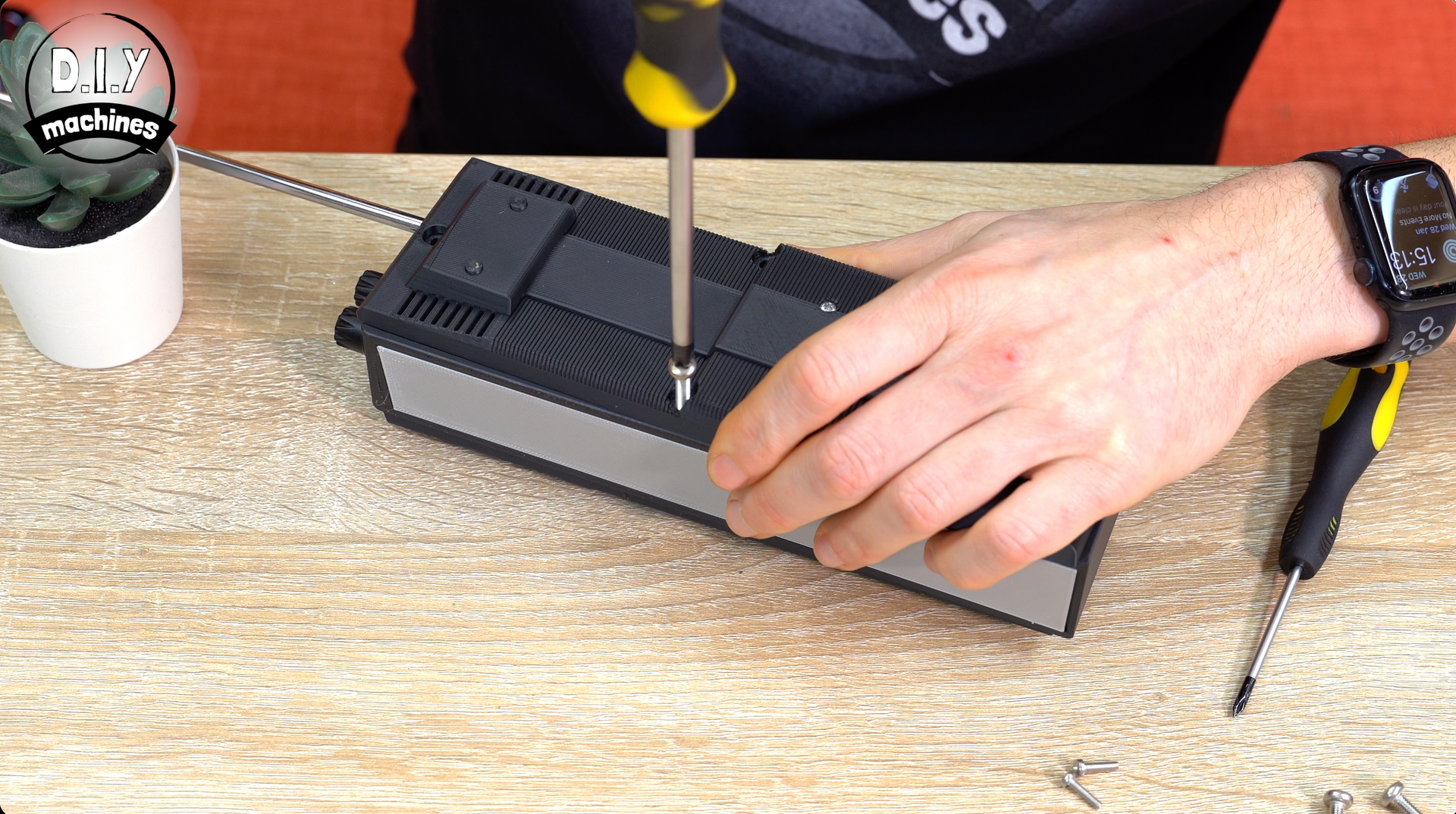 A person uses a screwdriver to remove a screw from the top of a black and silver rectangular electronic device on a table.