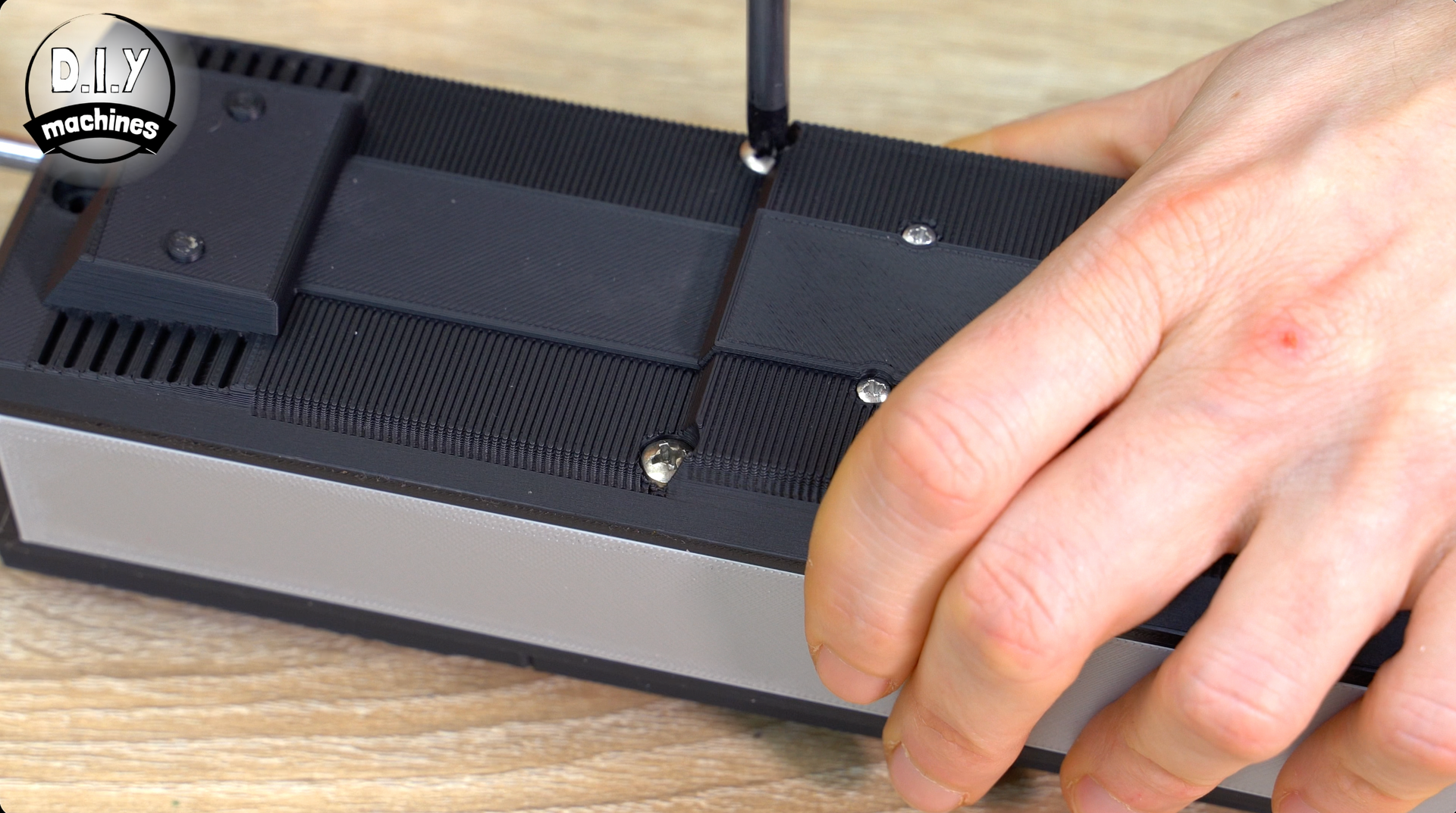 A hand uses a screwdriver to tighten a screw on the black textured underside of a metallic rectangular device.