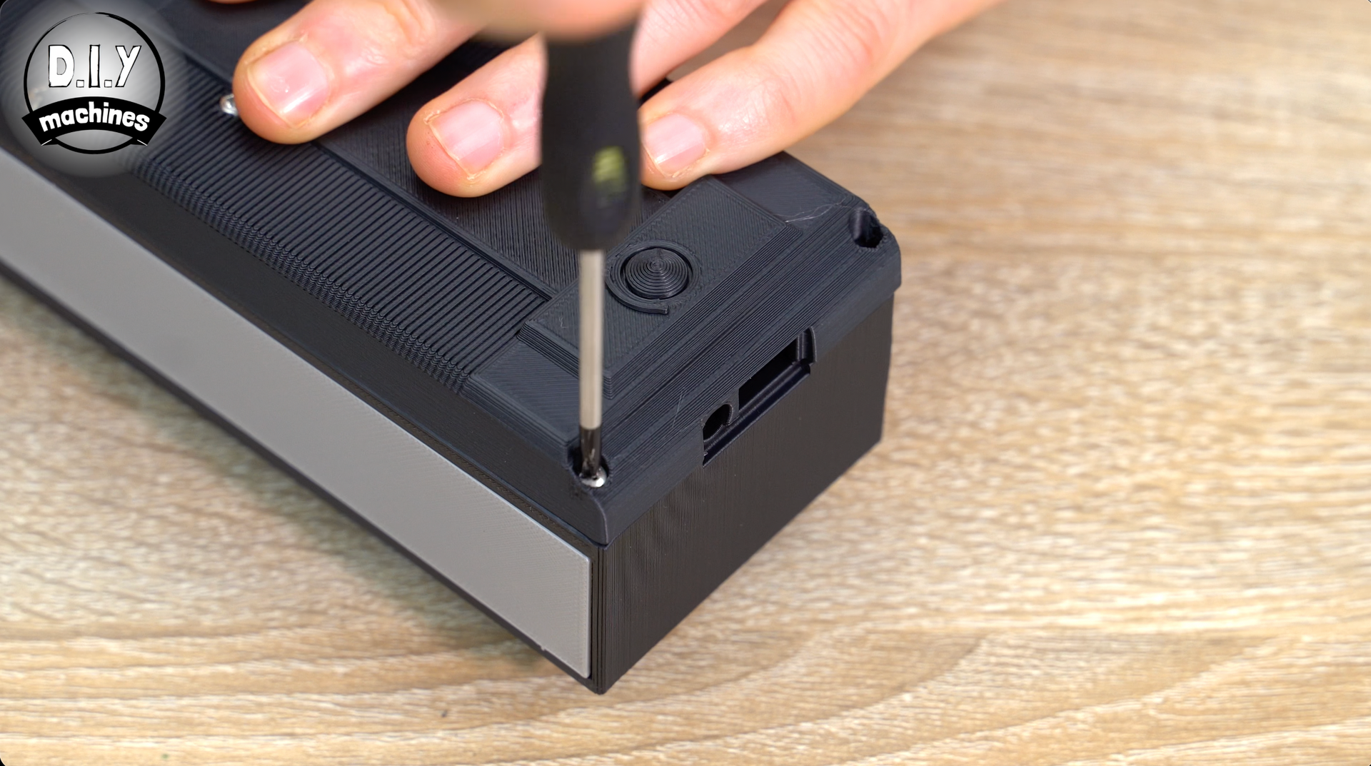 A hand uses a screwdriver to tighten a screw on the corner of a black, textured plastic rectangular device.