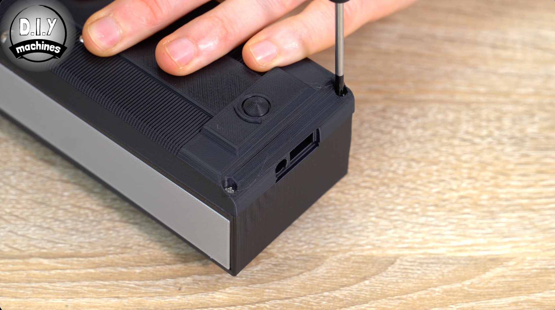A hand uses a screwdriver to tighten a screw on the corner of a black, textured 3D-printed case on a wooden surface.