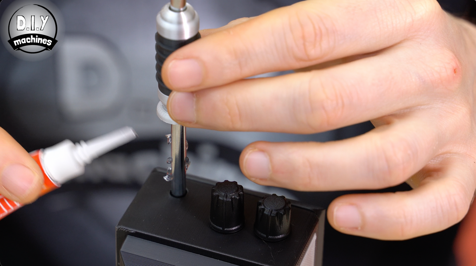 Hands use a screwdriver on a black electronics device while holding a small tube of glue nearby.