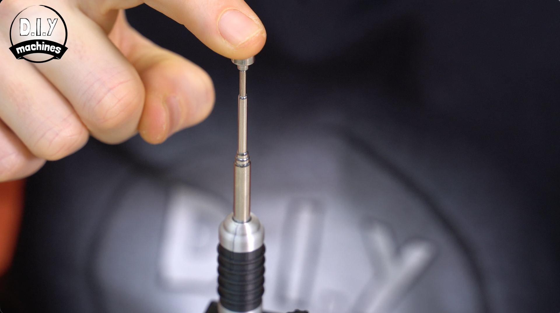 A hand holding a thin metal pin above a specialized cylindrical tool, with a blurred