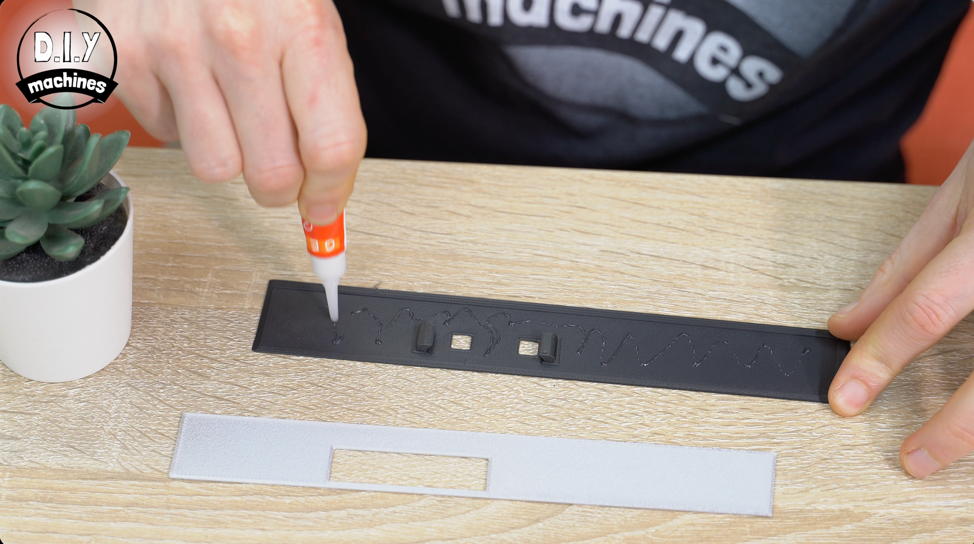Hands apply glue to a black component on a wooden desk, with a small succulent and a gray rectangular piece nearby.