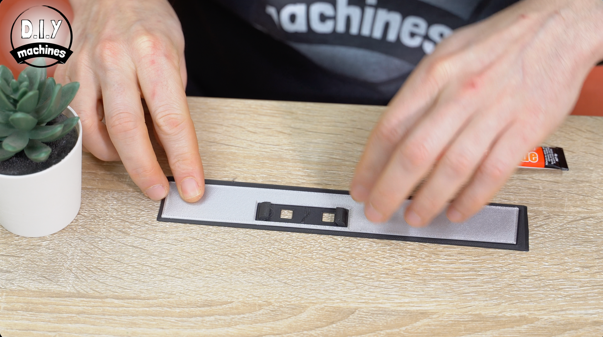 Hands smooth a strip of white, adhesive-backed tape onto a wooden surface, next to a small potted succulent.