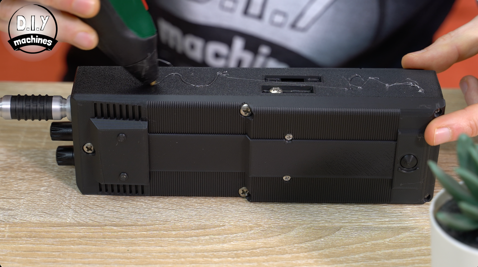 A person uses a hot glue gun on the textured, black surface of an electronic device on a wooden table.