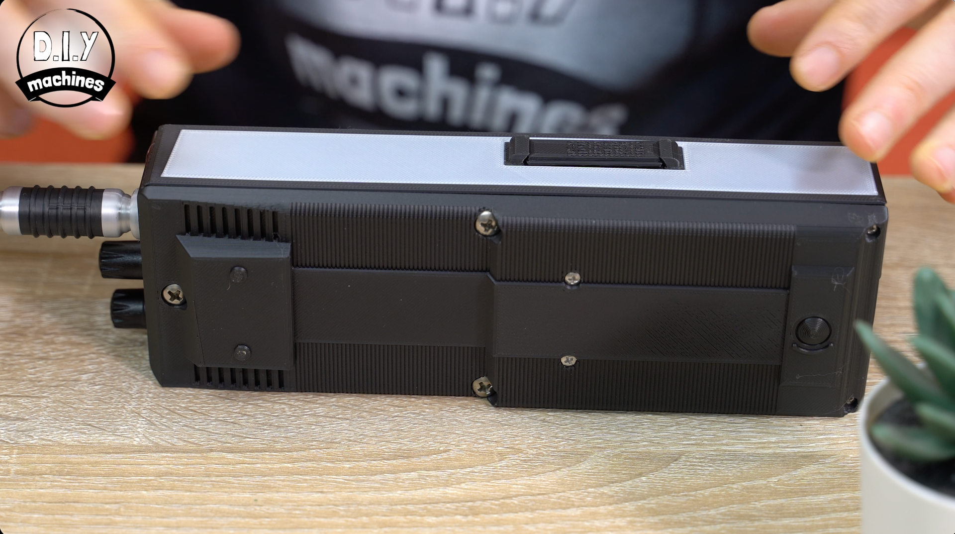 A black, rectangular electronic device with an antenna lies on a wooden surface, held by someone with visible hands.