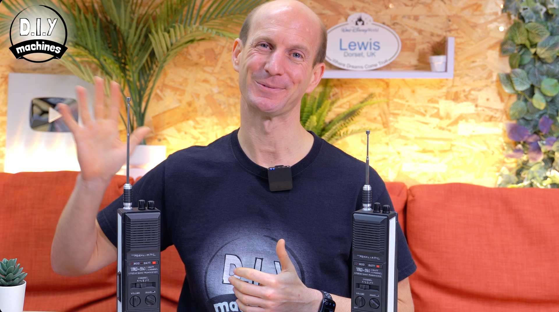 A person sits in front of an orange sofa with two walkie-talkies, waving at the camera in a room with plants.