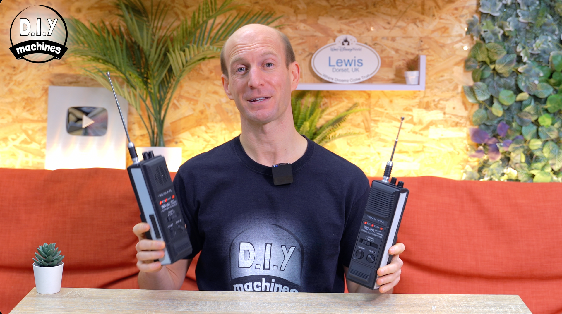 A person holds two black radio transceivers while sitting on an orange couch in a room with plants.