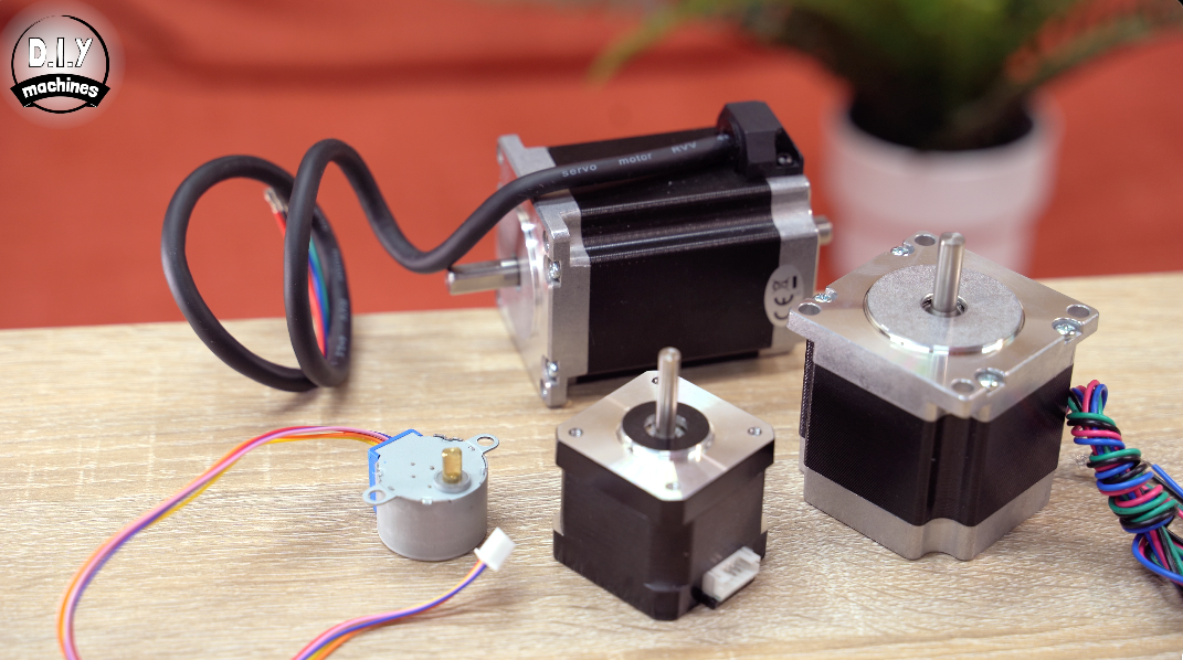 A collection of various sized stepper motors placed on a wooden surface with a small plant in the background.