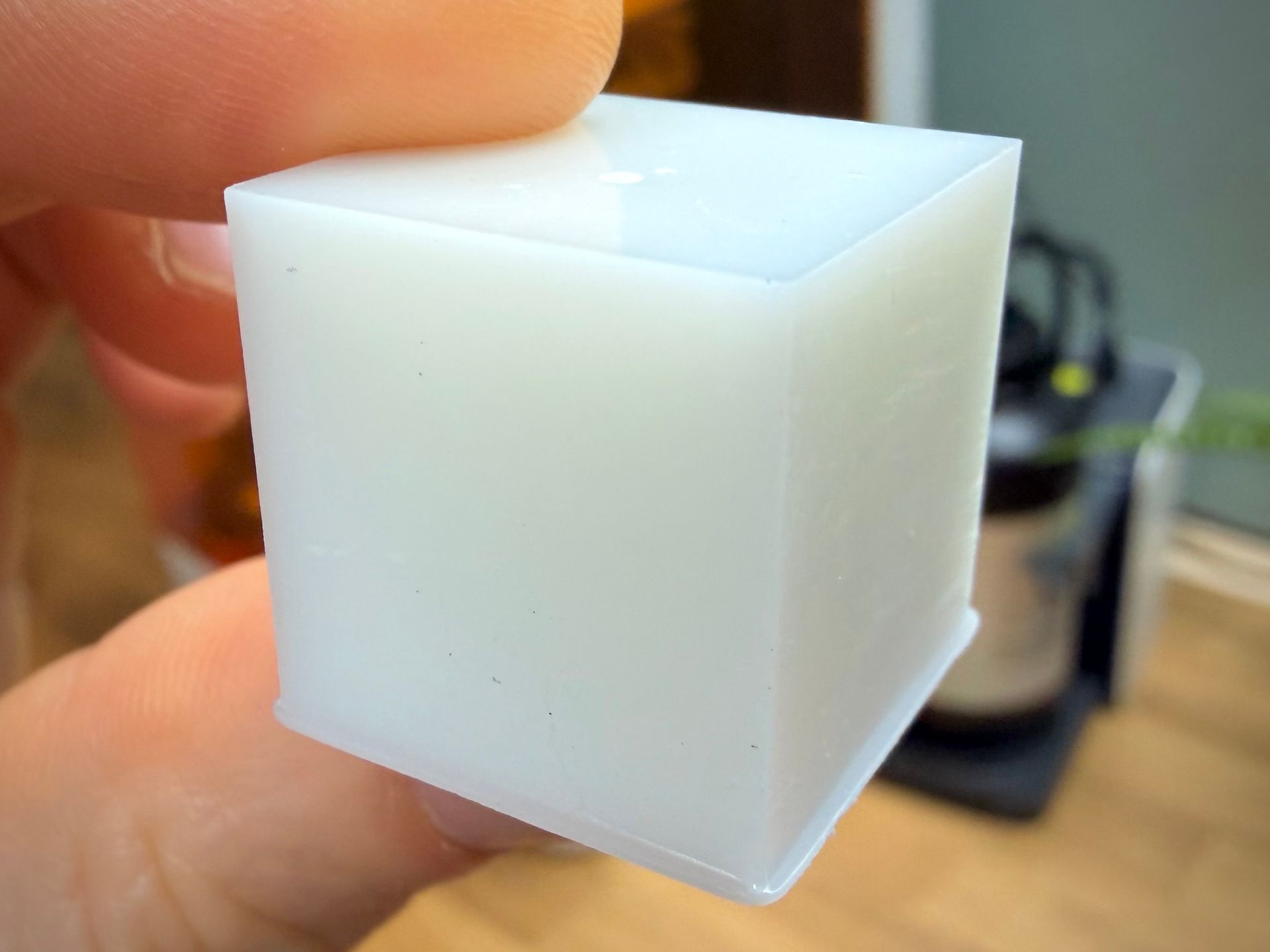 A hand holds a small, white, semi-translucent 3D-printed cube with a slight brim at the base.