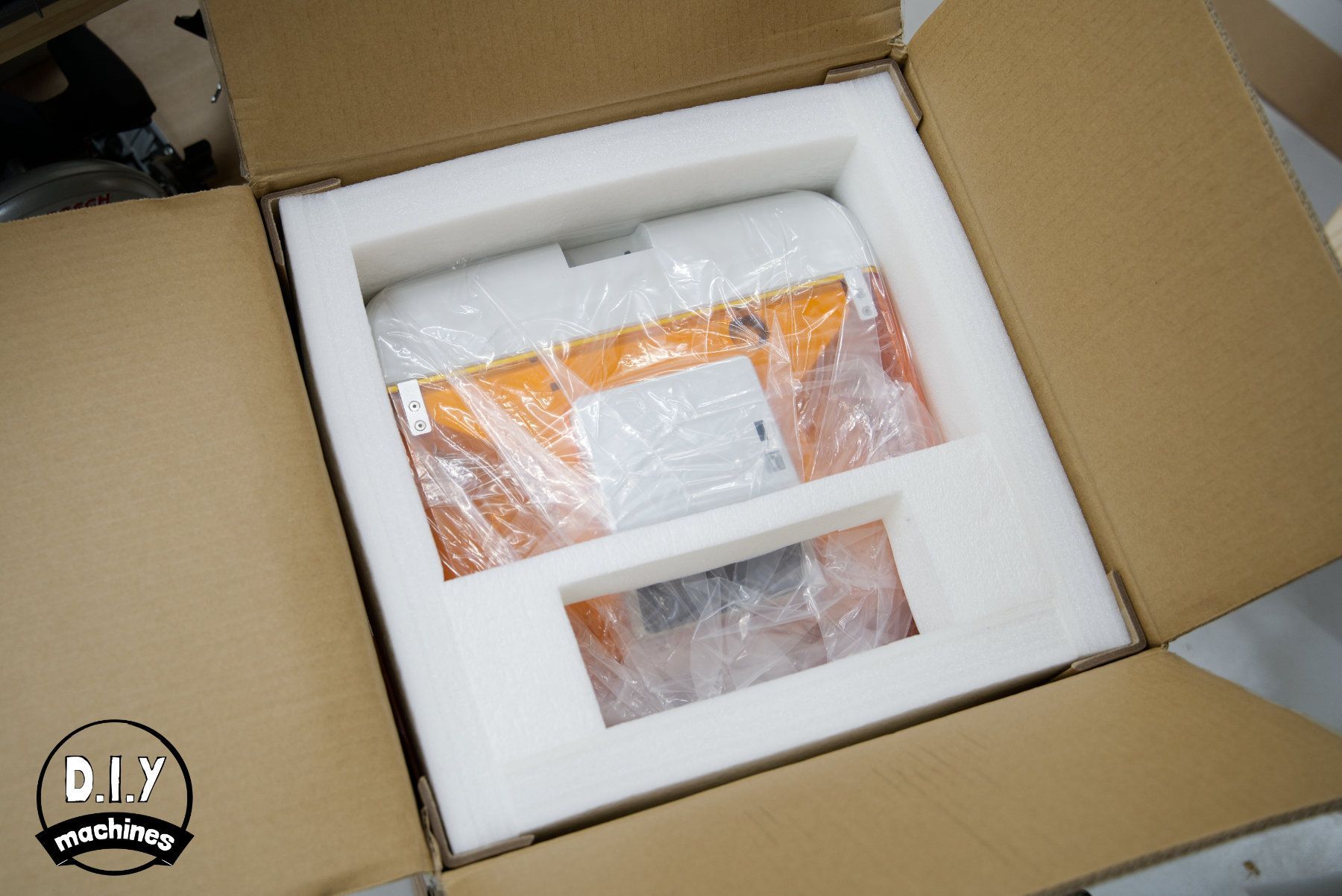 An orange and white device wrapped in clear plastic sits inside protective white foam packaging within a cardboard box.