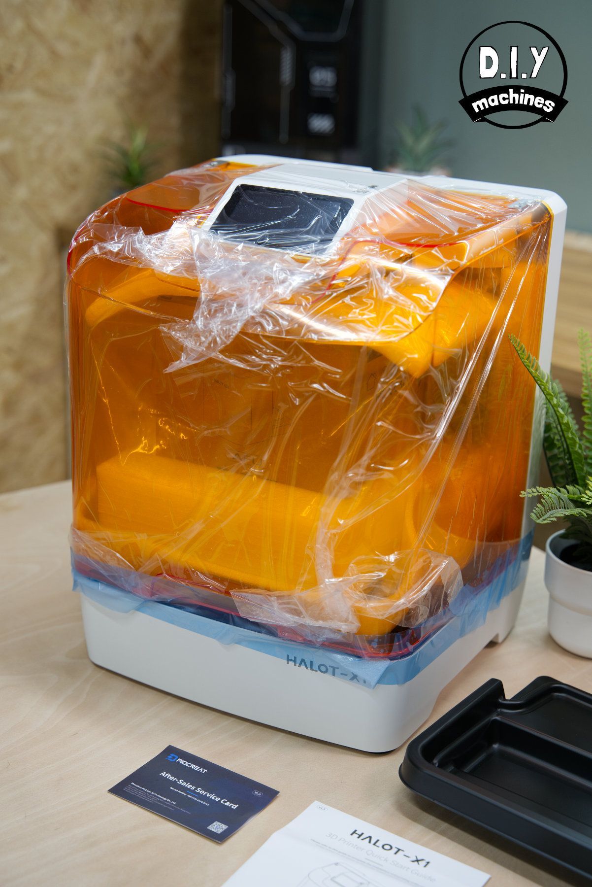 A brand new 3D resin printer wrapped in protective plastic sits on a desk next to its manual and a small potted plant.